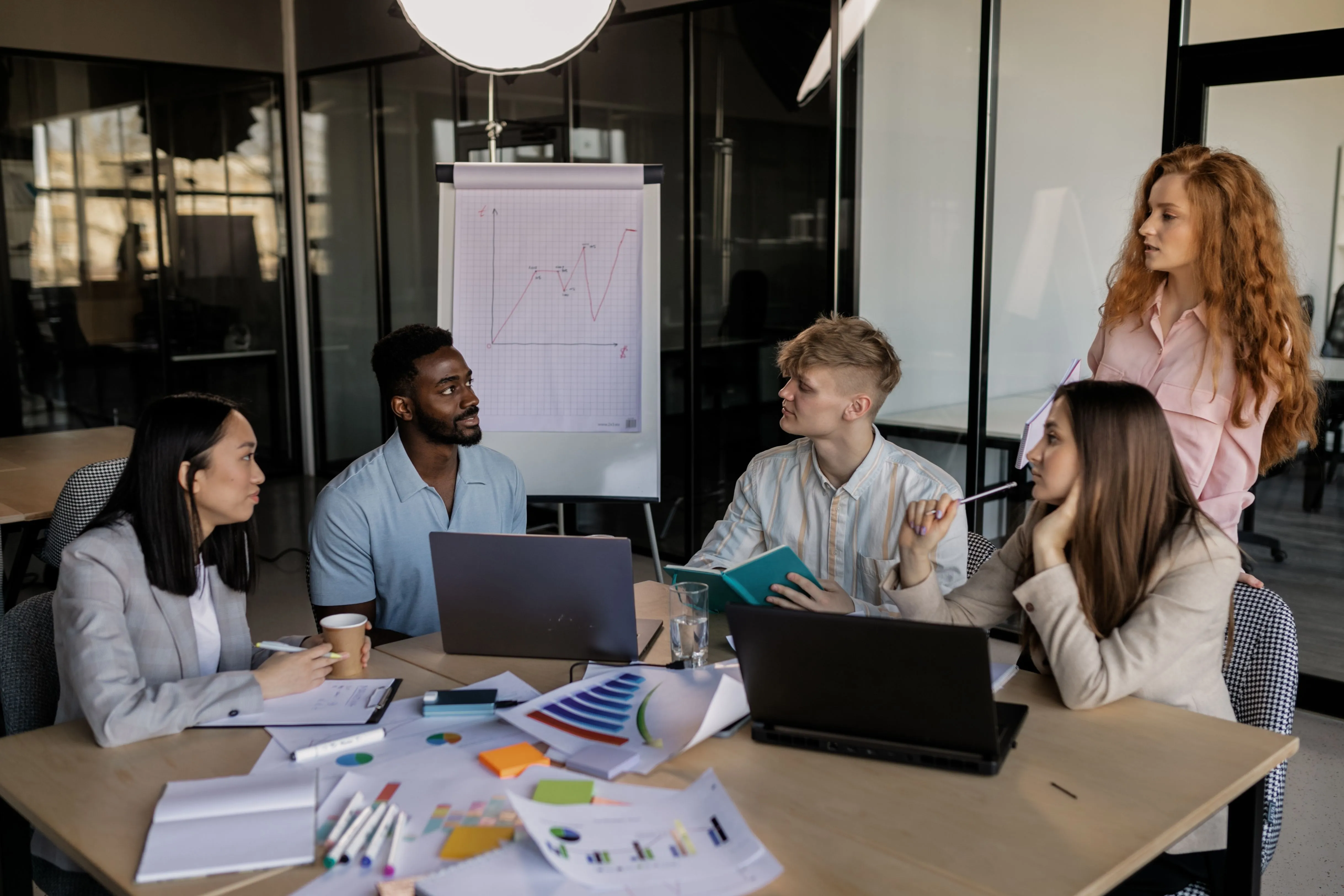 A diverse team collaborating around a table with laptops and printed charts