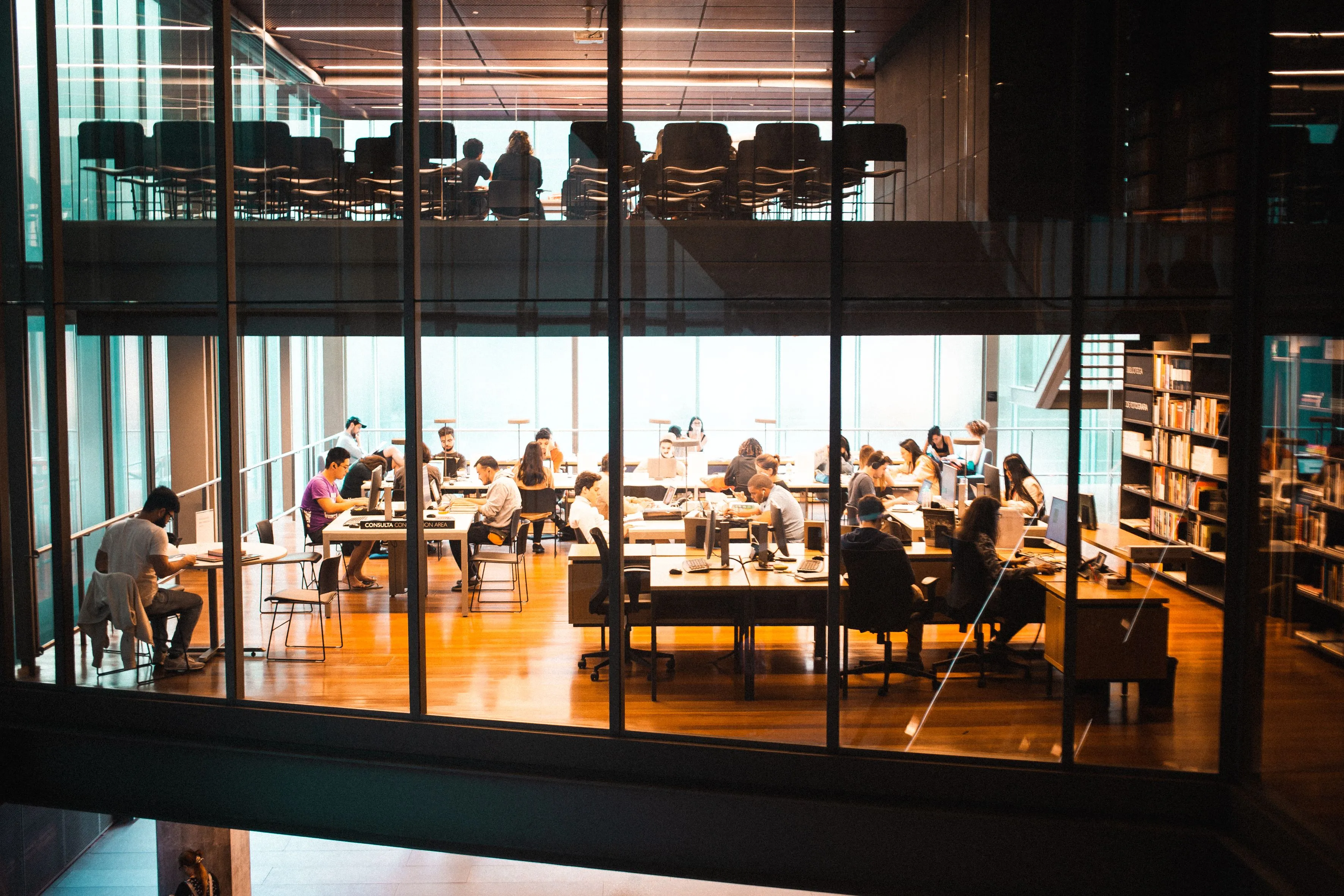 People working at desks across a busy open study hall lined with bookshelves